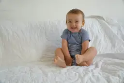 Lächelndes Baby im gestreiften Body sitzt auf weißer Decke.