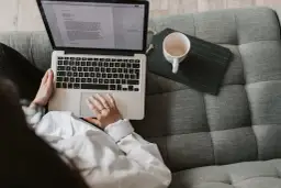 Scenarzysta pracuje nad nowym tekstem na laptopie, siedząc wygodnie na kanapie. Obok leży pusta filiżanka.