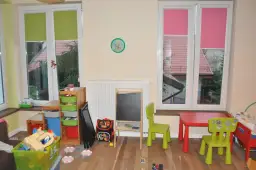 Pokój dziecięcy z zabawkami, stolikiem i krzesełkami. Okna z zielonymi i różowymi roletami, a za nimi widać dachy domów. Trofast inspiracje dla małych artystów.