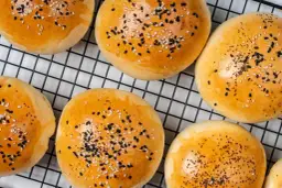 Złociste bułeczki do mini burgerów posypane sezamem i czarnuszką, na czarnej kratce.