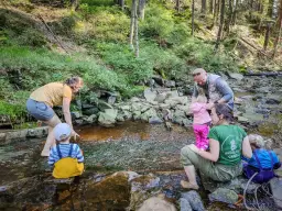 Rodzina z dziećmi pluska się w strumieniu. Dziecko w żółtym ogrodniczkach siedzi na kamieniu, a dorośli pomagają maluchom w wodzie. To piękny dzień na Baraniej Górze.