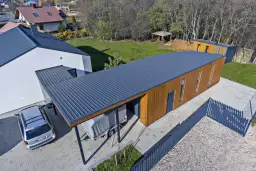 Nowoczesny budynek z drewnianą elewacją i ciemnym, **dachem jednospadowym**. Obok stoi samochód i przyczepa pod wiatą.