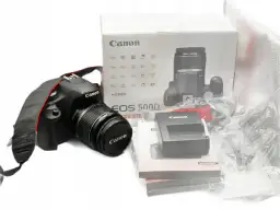 Aparat Canon EOS 500D z obiektywem i akcesoriami na białym tle.