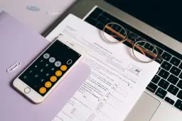 Telefon z kalkulatorem, okulary i dokumenty podatkowe na laptopie.