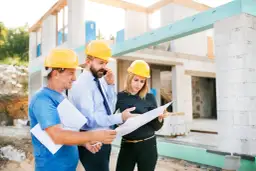 Trois personnes casquées discutent des plans sur un chantier. L'un d'eux est au téléphone, peut-être pour résoudre un litige travaux.
