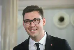 Uśmiechnięty Konrad Frysztak w okularach, w garniturze. Jego wykształcenie pozwala mu na pewność siebie w każdej sytuacji.