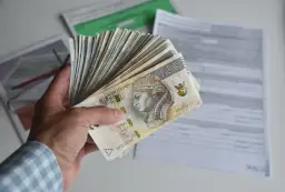 Dłoń trzyma plik banknotów 200 zł. W tle dokumenty, być może dotyczące tego, ile godzin to 0.5 etatu.