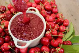 Cuchara vierte mermelada de cerezas casera en un cuenco blanco, rodeado de cerezas frescas y hojas verdes.