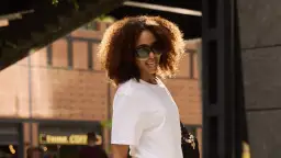 Frau mit lockigem Haar und Sonnenbrille trägt ein weißes T-Shirt.