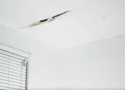 Widoczny pęknięcie na suficie w bloku, odsłaniające fragmenty konstrukcji. Obok okno z białymi żaluzjami.