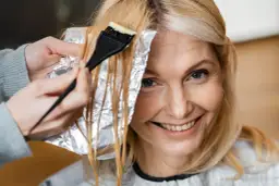 Kobieta z uśmiechem na twarzy farbuje włosy. Jasny blond może postarzać, ale ona wygląda promiennie.