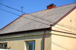 Dach pokryty falistym eternitem, z ceglanym kominem i liniami energetycznymi przecinającymi błękitne niebo.