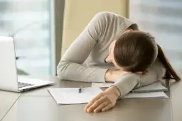Kobieta z opuszczoną głową na ramieniu przy biurku z laptopem i dokumentami.