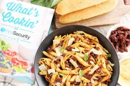 Makaron penne z suszonymi pomidorami, parmezanem i bazylią w czarnej misce, obok pieczywa i książki kucharskiej.