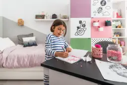 Dziewczynka rysuje w swoim małym pokoju dla dziecka. Na biurku leżą kolorowe kamyczki i papier.