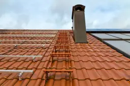 Pomarańczowe stopnie kominiarskie do dachówki prowadzą do komina na dachu z panelami fotowoltaicznymi.