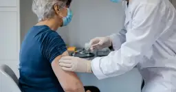 Starsza pani otrzymuje szczepionkę na covid. Lekarz w białym fartuchu i rękawiczkach podaje zastrzyk.