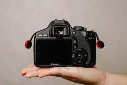Dłoń trzyma czarną lustrzankę Canon z czerwonymi elementami smyczy.