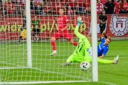 Napięta akcja w polu karnym podczas meczu Widzew Łódź – Lech Poznań. Bramkarz w zielonym stroju broni strzał, a zawodnik w niebieskiej koszulce leży na murawie.
