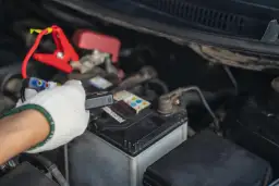 Ręka w białej rękawiczce podłącza prostownik do akumulatora. Wreszcie, kiedy prostownik pokazuje, że akumulator jest naładowany, można ruszać.