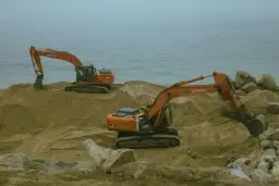 Dwie koparki pracują na piaszczystym brzegu. Widoczny jest element roboczy koparki, gotowy do pracy.