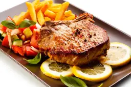 Chuletas de cerdo al horno jugosas, acompañadas de papas fritas crujientes, ensalada fresca y rodajas de limón.