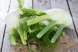 Kilka świeżych bulw fenkułu z zielonymi łodygami i liśćmi, ułożonych na drewnianym blacie. Fenkuł co to? To aromatyczne warzywo.