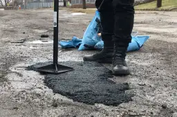 Pracownik ubija asfalt na zimno, naprawiając dziurę w drodze. Obok leży niebieski worek z materiałem.