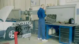 Mechanik w niebieskim kombinezonie przy silniku samochodu. Obok skrzynka z narzędziami, płyny i kanistry. Unikaj mandatu za brak przeglądu, dbaj o auto.