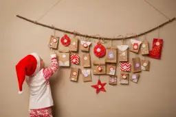 Dziecko w czapce Mikołaja wskazuje na zawieszone na gałęzi paczuszki z numerami. To inspiracja, jak zrobić kalendarz adwentowy z kartonu.