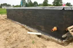 Pomarańczowa rura kanalizacyjna przechodzi przez fundament z czarnej cegły, przygotowywany do dalszych prac budowlanych.