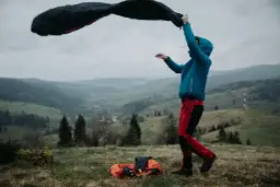 Osoba w niebieskiej kurtce i czerwonych spodniach rozkłada śpiwór na tle gór. Warto wiedzieć, jaki śpiwór wybrać na taką wyprawę.