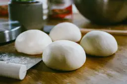 Cztery kulki surowego ciasta na pizzę leżą na drewnianym blacie obok noża.