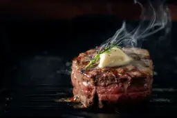 Soczysty stek wołowy z masłem i rozmarynem na grillu, z którego unosi się para.