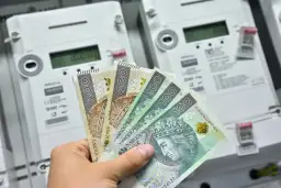 Dłoń trzyma polskie banknoty przed licznikami energii elektrycznej. Zastanawiasz się, ile prądu zużywa zamrażarka miesięcznie?