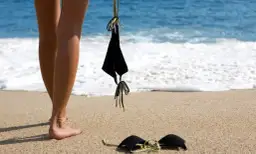 Nogi na plaży, obok leżą czarne bikini. Słońce grzeje, a fale obmywają brzeg. Czas na kąpiel dla golasy.