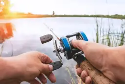 Dłonie trzymające niebiesko-srebrną wędkę spinningową nad wodą.