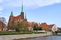Ostrów Tumski we Wrocławiu z katedrą i zielonymi wieżami, nad rzeką, z ludźmi spacerującymi po bulwarze.
