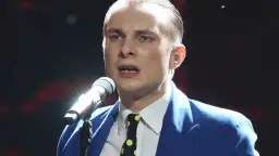 Ralph Kaminski, w niebieskiej marynarce i krawacie w grochy, śpiewa do mikrofonu. Jego partner, wokalista, jest w centrum uwagi.