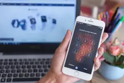 Eine Hand hält ein iPhone mit gesperrtem Bildschirm und Ziffernfeld. Im Hintergrund ein Laptop.
