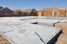 Fundament domu z chudego betonu, z widoczną konstrukcją szkieletową przyszłego budynku i rurami instalacyjnymi.