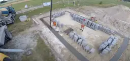 Pracownicy wylewają beton na fundamenty. Widać koparkę, betoniarkę i sterty pustaków.