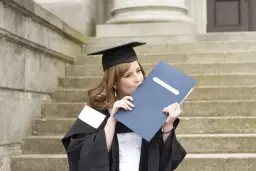 Absolwentka studiów prawniczych całuje dyplom, ubrana w togę i biret, na tle schodów.