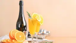 Dwa kieliszki orzeźwiającej mimosa z plasterkami pomarańczy i gałązką rozmarynu, obok butelki szampana i shakera.