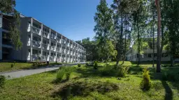 Hotel NAT w Krynicy Morskiej otoczony zielenią, z długim budynkiem o wielu balkonach i drzewami w tle.