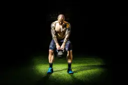 Mężczyzna z tatuażami trzyma kettlebell na tle ciemności i zielonej trawy.