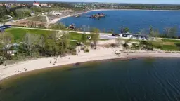 Słoneczny dzień nad zalewem Mietkowskim. Ludzie spacerują po piaszczystej plaży, a w oddali widać budynki i sprzęt pływający.
