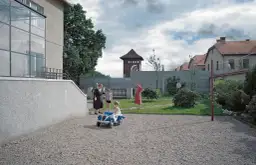 Strefa interesów recenzja: rodzina w ogrodzie, za nią mur, druty kolczaste i wieża strażnicza. Dzieci bawią się autem.