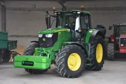 Zielony John Deere 6195M, z kierowcą w kabinie, stoi obok przyczepy. Czy to najlepsza marka ciągnika?