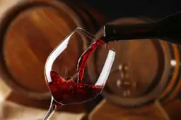 Czerwone wino, bogate w taniny, leje się z butelki do kieliszka, na tle drewnianych beczek.